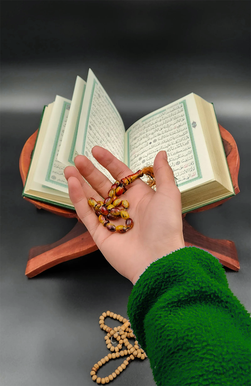Home A hand holding prayer beads beside an open Quran, symbolizing spirituality and devotion.